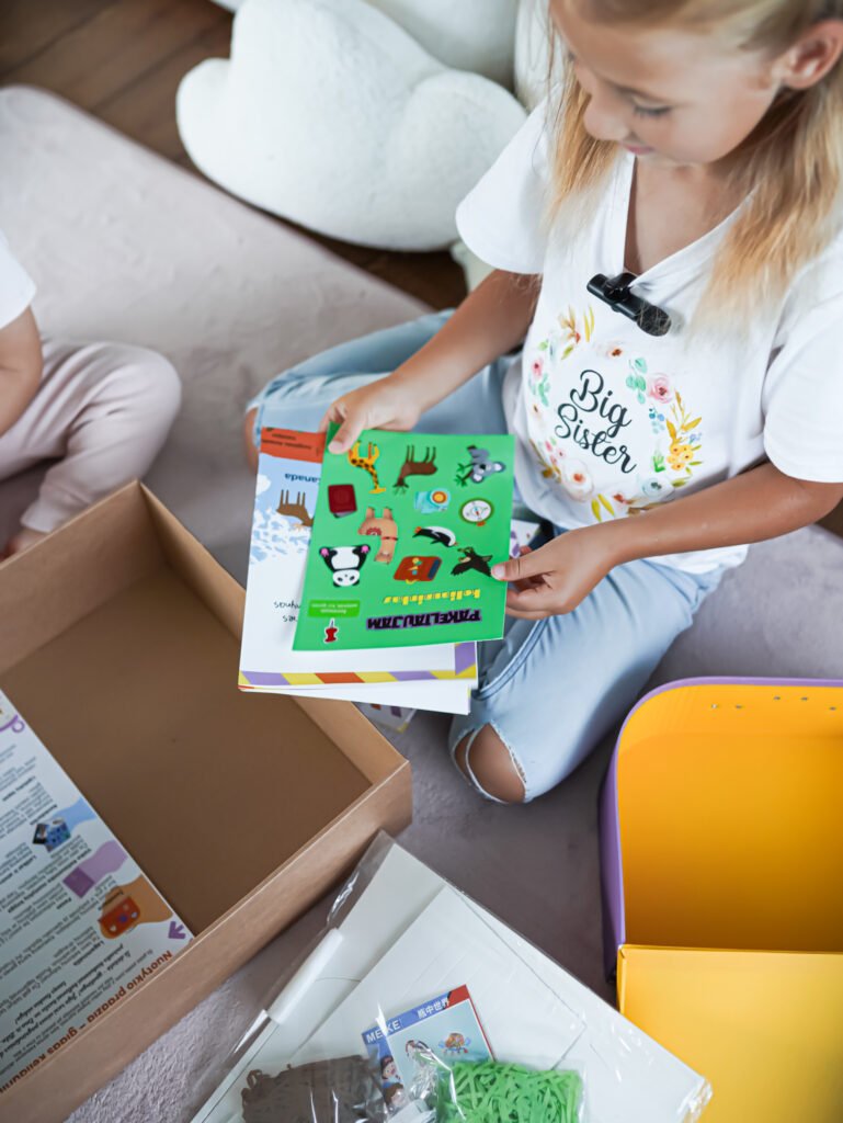Young girl sits on the floor reading a bright sticker/activity book taken from a cardboard box.