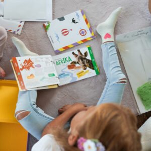 Two kids sit on a beige carpet and read colorful children's books laid out on the floor.