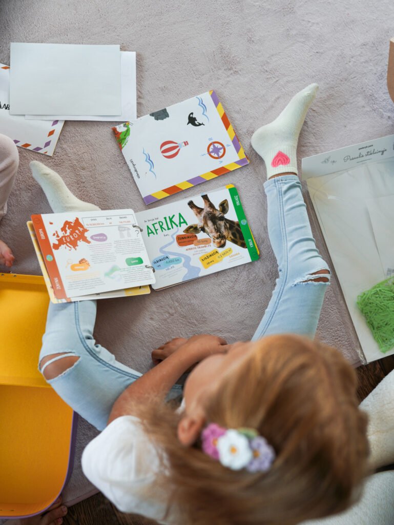 Two kids sit on a beige carpet and read colorful children's books laid out on the floor.