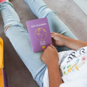 Person seated on a rug holds a purple passport-style booklet with a globe illustration on the cover while wearing ripped light-blue jeans.