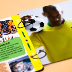 Blurred person in a yellow shirt holds a colorful magazine spread featuring a soccer ball on a peach background.