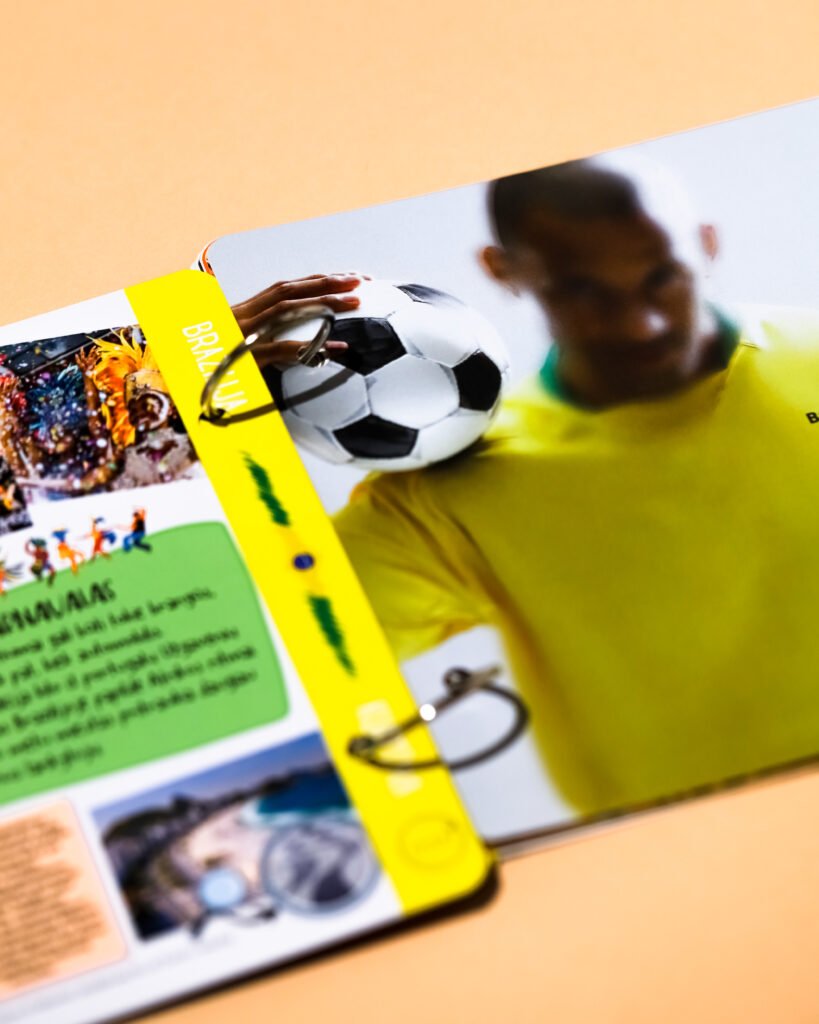 Blurred person in a yellow shirt holds a colorful magazine spread featuring a soccer ball on a peach background.
