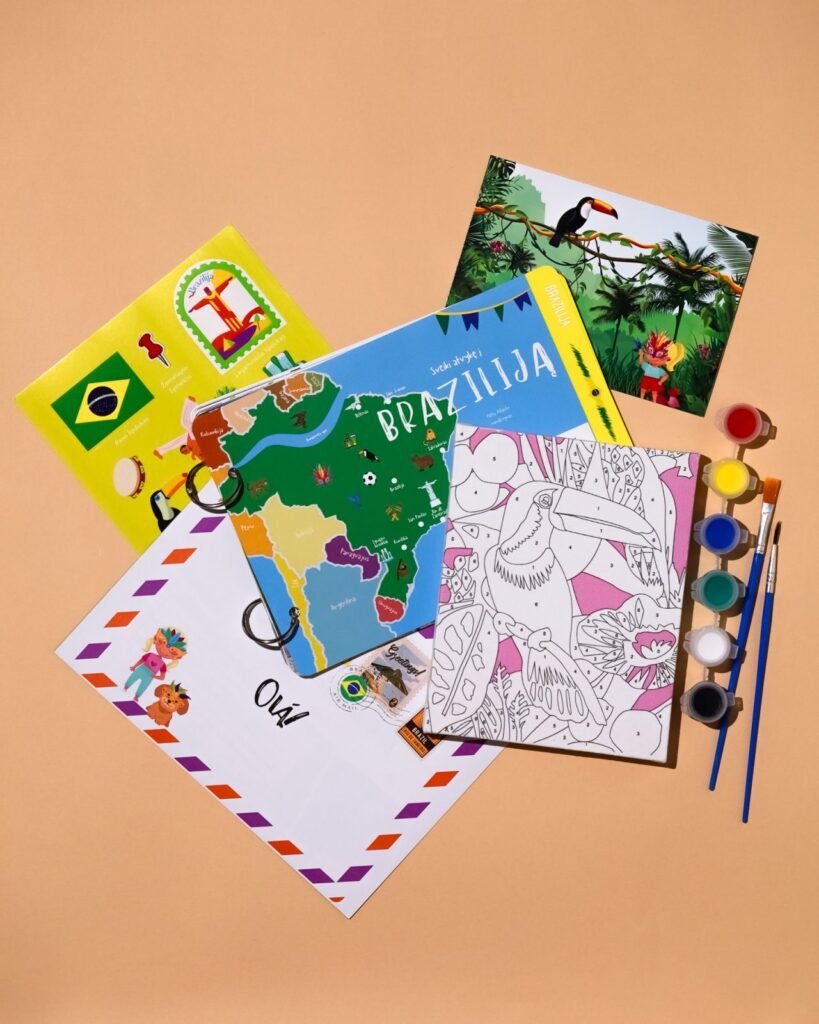 Flat lay of colorful travel books, a Brazil map, a coloring page, and watercolor paints with brushes on a peach background.