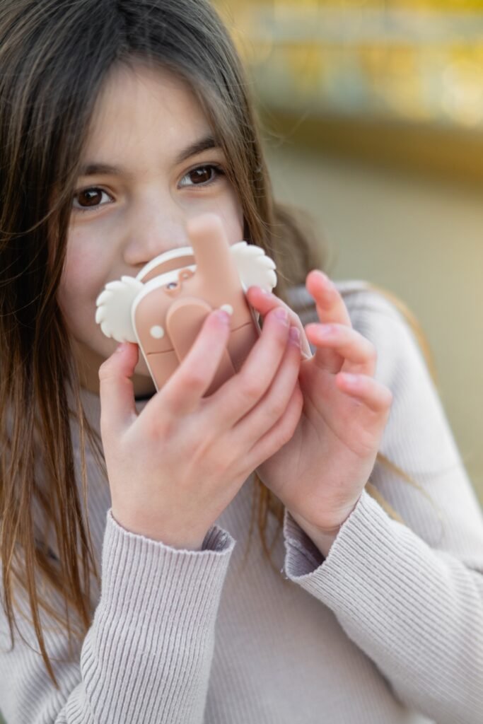 Young girl with long hair holding a beige plastic device to her nose, the object has white wing-like parts on the sides and she looks slightly to the side in a soft outdoor setting.