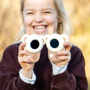 Young girl with blonde hair smiling broadly while holding two pink toy cameras toward the viewer with round screens and faux lenses, outdoors.