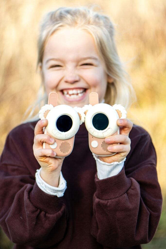 Young girl with blonde hair smiling broadly while holding two pink toy cameras toward the viewer with round screens and faux lenses, outdoors.