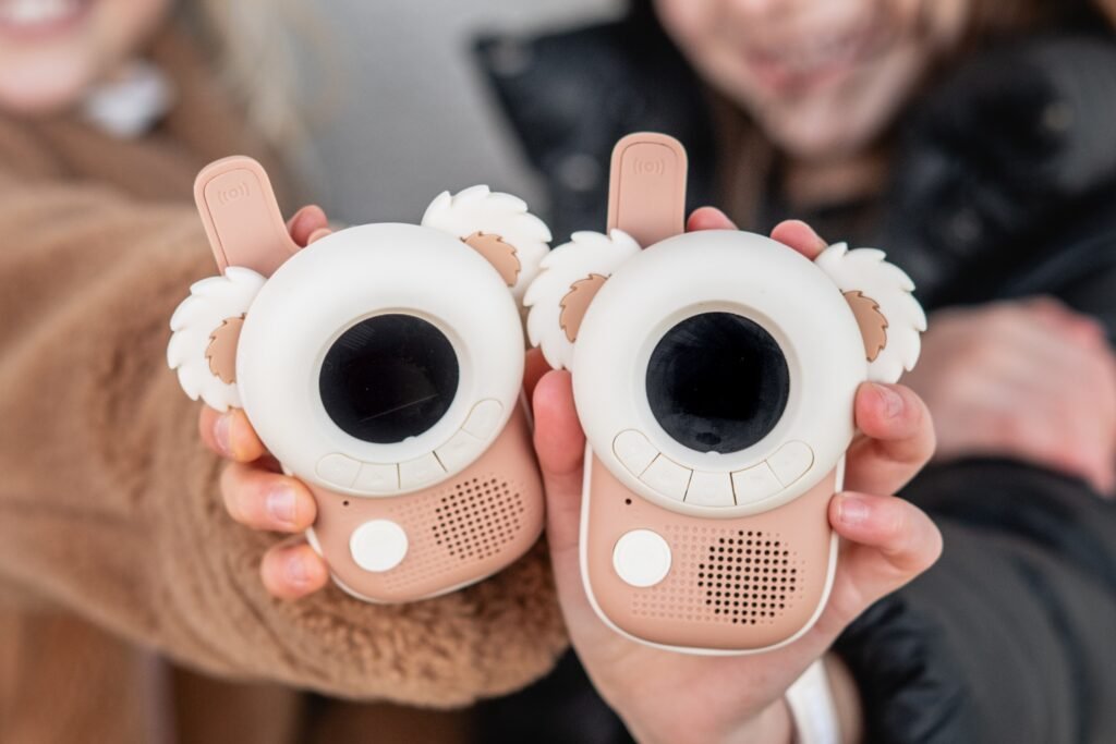 Two cartoon-like toy cameras held up for a photo, each with a large circular eye lens and fluffy ears, in pink and white.