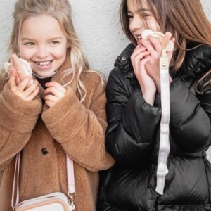 Two smiling girls in warm coats holding white fluffy earmuffs against a light wall.