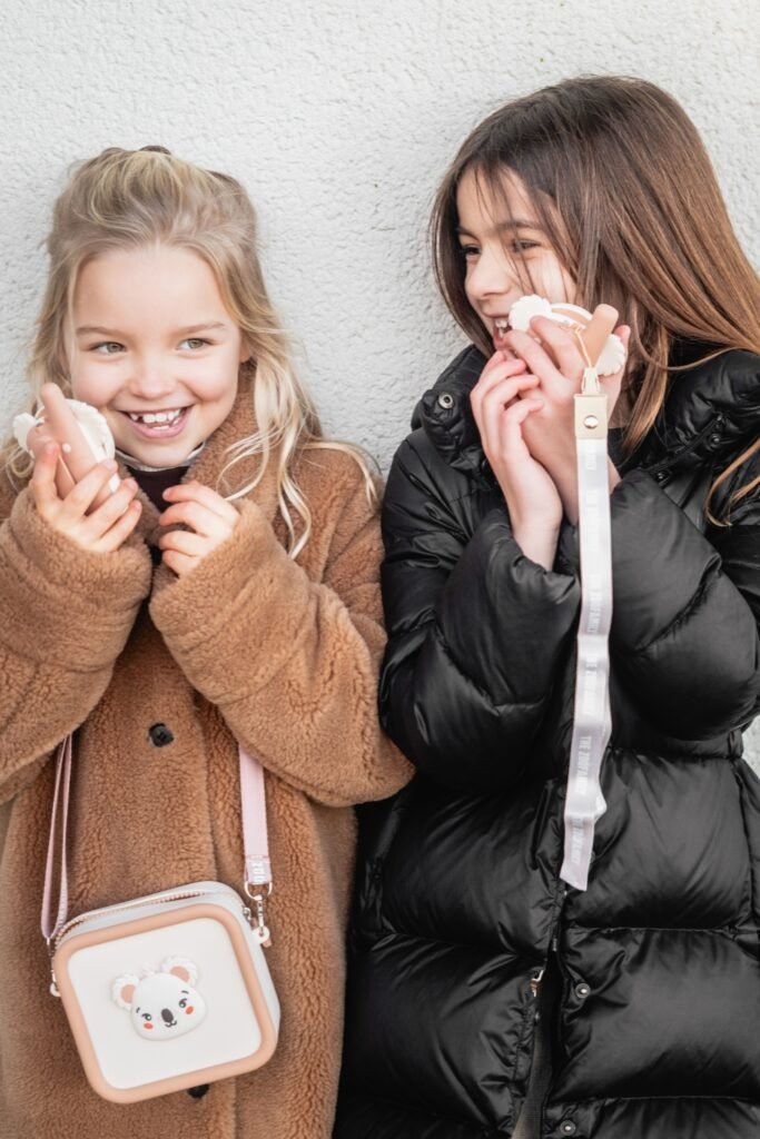 Two smiling girls in warm coats holding white fluffy earmuffs against a light wall.