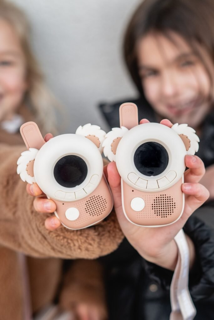 Two pink bear-themed toy cameras with round lens screens and fluffy ears, held by smiling kids behind them back-to-back in a close-up.