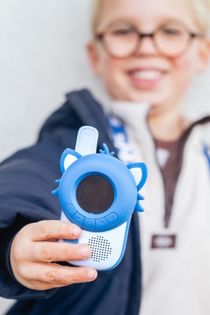 A child wearing glasses holding up a blue handheld kids' toy with a round display and cat-ear accents toward the camera.