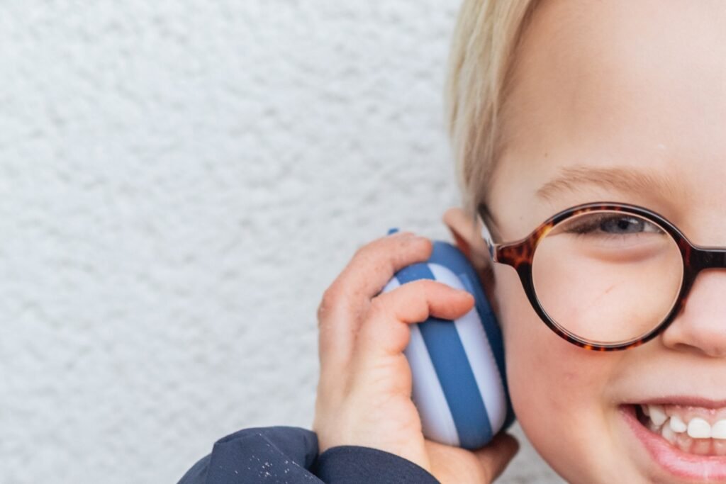 Close-up of a smiling child with tortoiseshell glasses wearing blue and white over-ear headphones against a light wall surface.