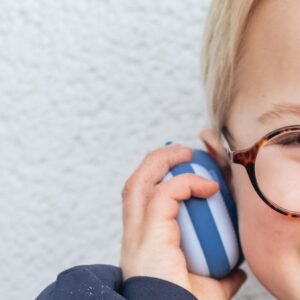 Close-up of a smiling child with tortoiseshell glasses wearing blue and white over-ear headphones against a light wall surface.