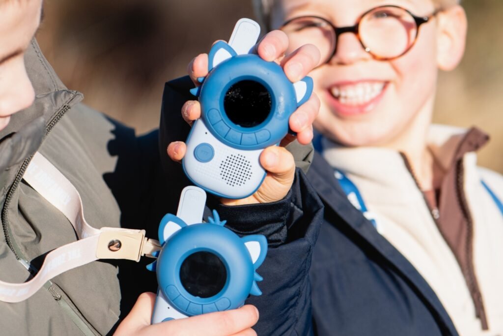 Two children outdoors proudly display blue, round-screen toy devices with white ear-like accents, smiling at the camera.