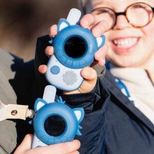Two children outdoors proudly display blue, round-screen toy devices with white ear-like accents, smiling at the camera.