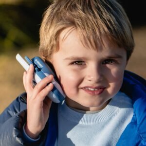 Young boy wearing a light blue sweater and blue jacket holds a blue walkie-talkie to his ear outdoors, smiling in sunlight.