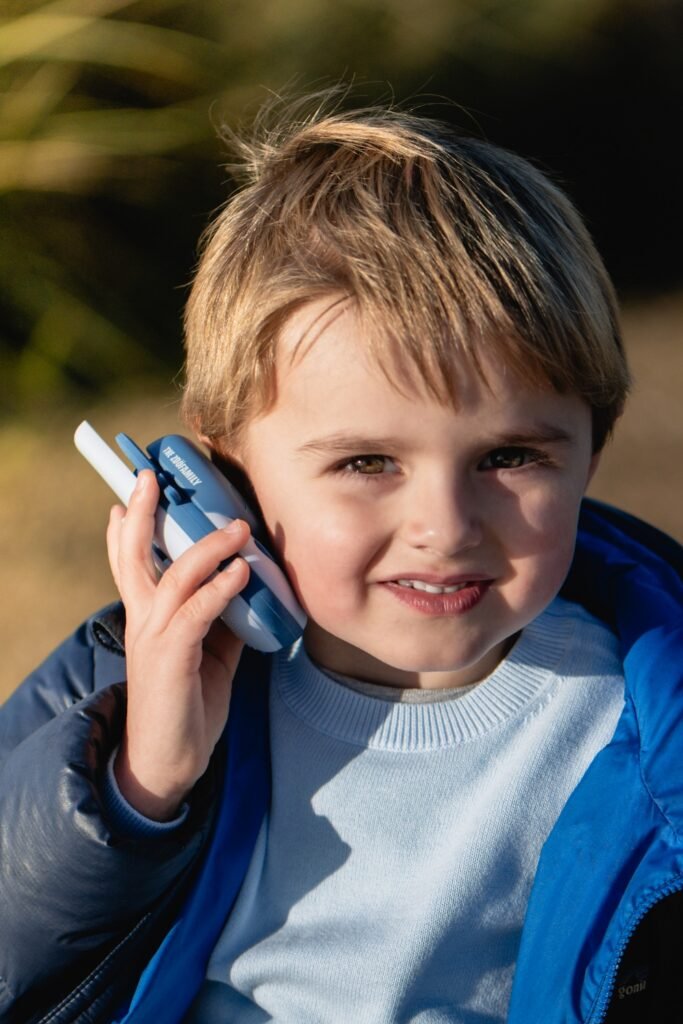 Young boy wearing a light blue sweater and blue jacket holds a blue walkie-talkie to his ear outdoors, smiling in sunlight.
