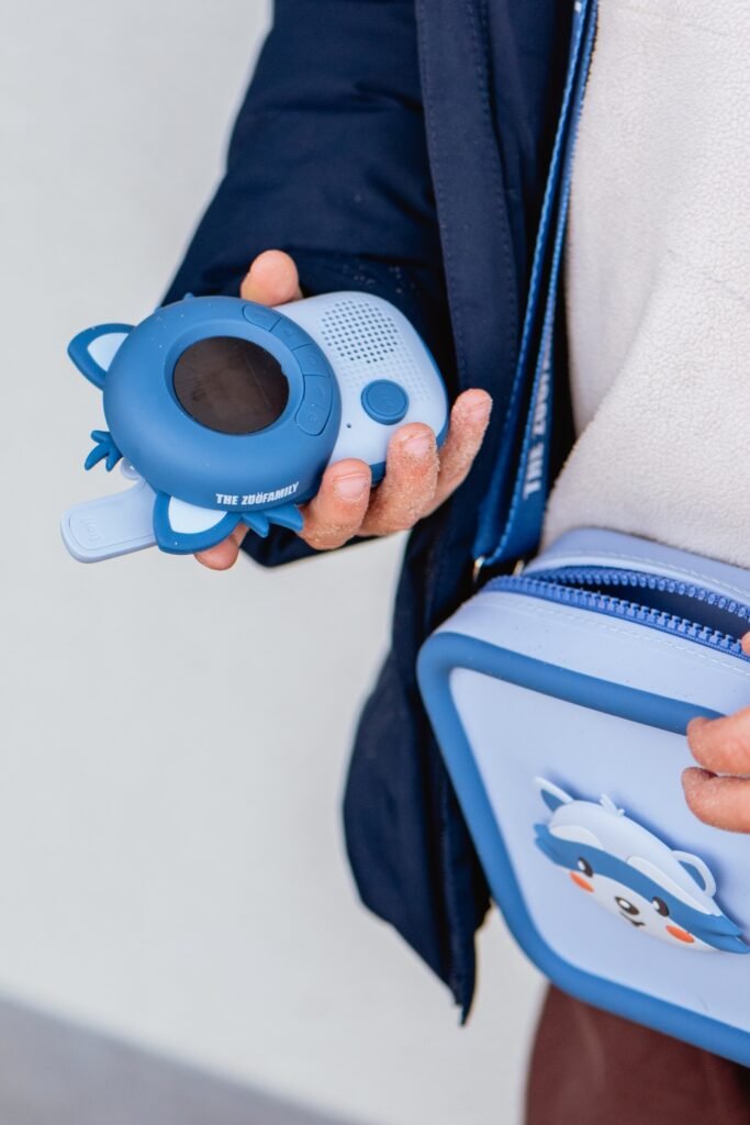 Child's hand holding a blue toy camera with a circular screen and buttons labeled The Zoo Family.