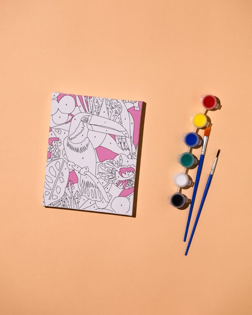 Open coloring book with a detailed line drawing of birds and flowers on a peach background, with paints and brushes to the right.